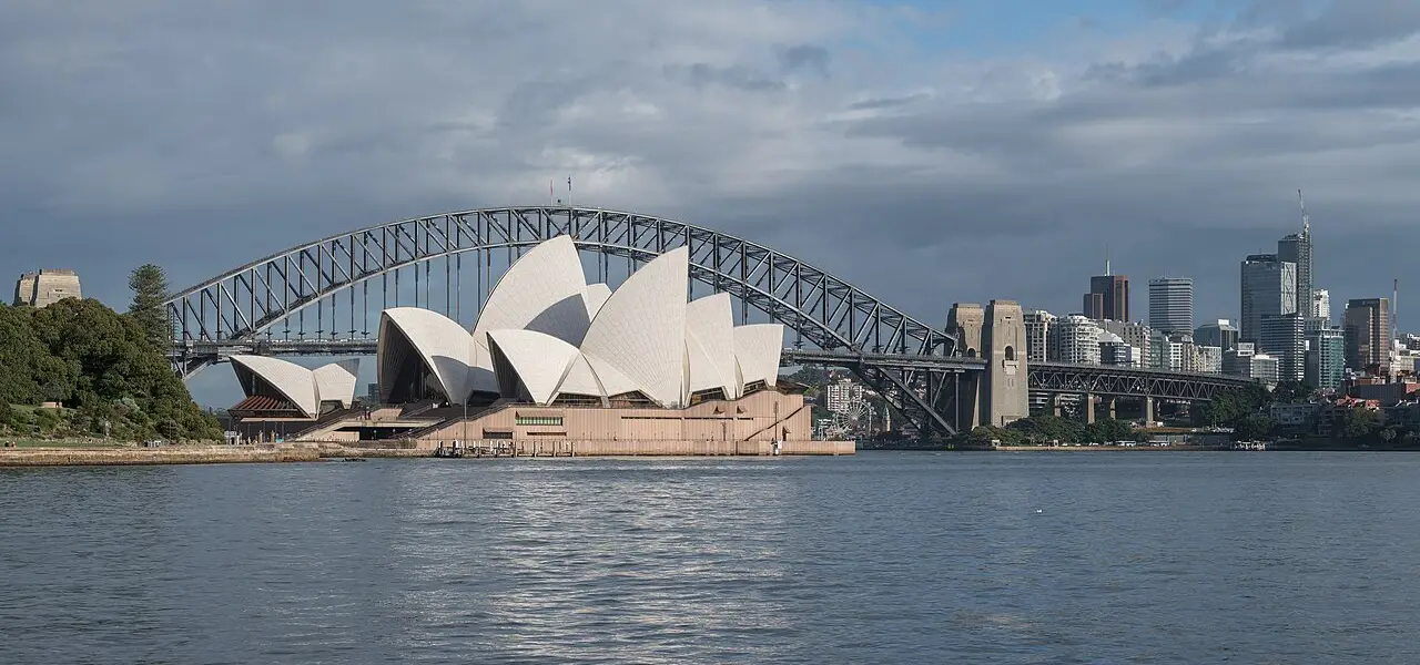 The Sydney Opera House and Sydney Harbour Bridge across the harbour water, with the city skyline on the right on a partly cloudy morning.